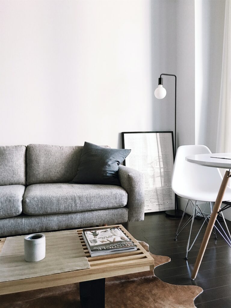 Image to emphasize the basics of neutral colors. A living room with white walls, grey couch, grey accent pillow, neutral wall art, black floor lamp, wooden coffee table, and a white dining table with white chair. 